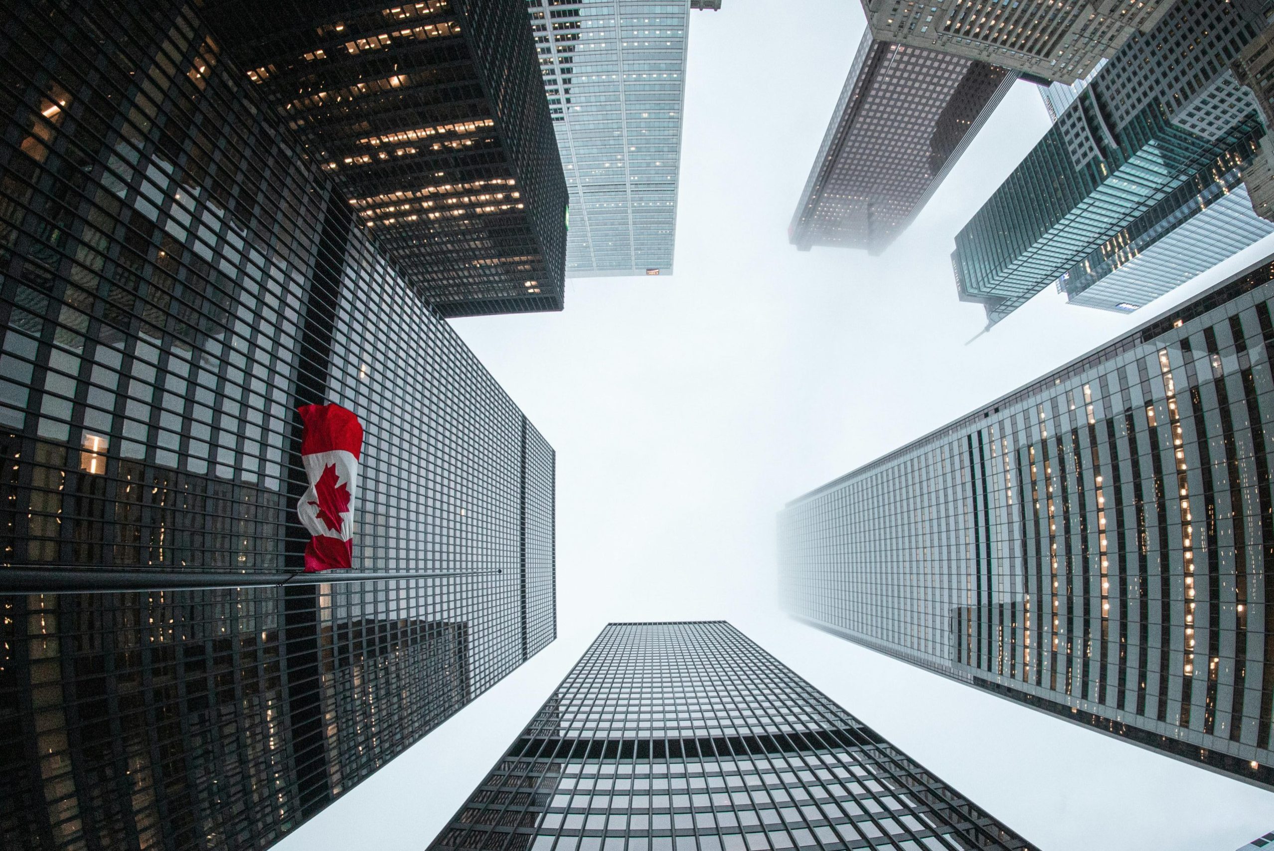 Looking up at Toronto skyscrapers with a Canadian flag on a cloudy day.